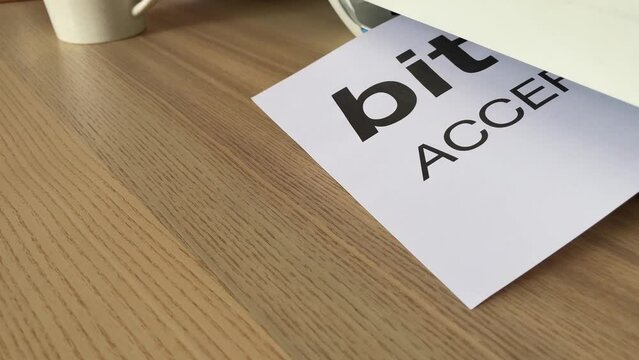 Printing &ldquo;bitcoin ACCEPTED HERE&rdquo; text written message on paper sheet document. Close up A4 page being printed from multifunction scanner printer on wooden desk in office. Hand take paper from printer