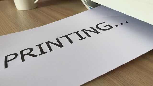 Printing &ldquo;PRINTING&rdquo; text written message word on paper sheet document. Close up A4 page being printed from multifunction scanner printer on wooden desk in office. Hand take paper from printer