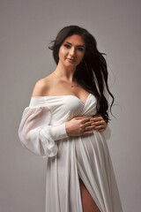 Pregnant brunette woman in white dress posing at camera isolated on grey background. Expecting baby