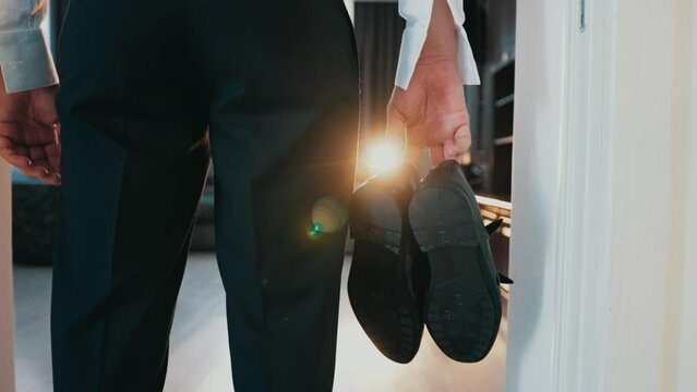 An elegantly dressed man stands in the doorway and holds a pair of shoes. Rear view. Businessman in pants and shirt with unbuttoned cuffs looks into the room. Flashlight light