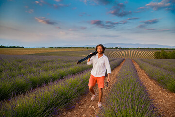 The handsome brutal man with long brunette hair is walking with tripod in the field of lavender in provence near Valensole, France, clear sunny weather, in a rows of lavender, red shorts, white shirts