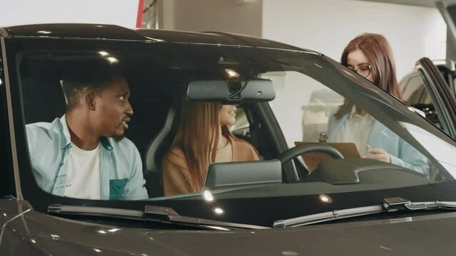 Female seller working with multiracial couple at modern auto center. Happy african man and caucasian woman consulting with manager while choosing new vehicle. Multi ethnic couple and saleswoman.
