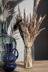 A composition of vases with dried flowers of a different type on a wooden table in light room interior with shadows. 