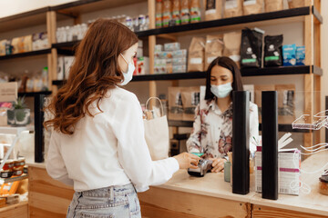Girl buyer in a mask makes a purchase in the store. Saleswoman or small business owner wearing medical mask at the counter in cafe or small shop.