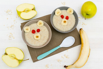 Funny cute kids childrens baby's healthy breakfast lunch oatmeal porridge in bowl look like bear face decorated with apple, banana, dried berry fruits. dessert food art on white wooden table