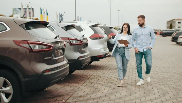 Saleswoman showing something on clipboard to male customer of dealership center. Happy bearded man buying modern car with sale banner on windshield. Female seller helping male client shoosing car.