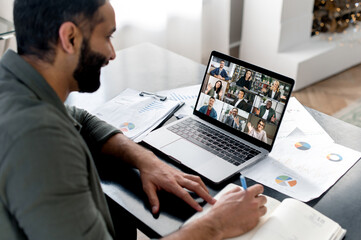 Fototapeta na wymiar Business briefing. Indian guy looking at laptop screen, talking on video call with multinational business group, holds conference, virtual meeting, brainstorming with colleagues online, takes notes