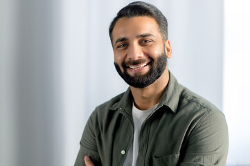 Obraz premium Portrait of indian or arabian positive successful businessman, manager, freelancer, office worker, wearing casual stylish shirt, stand in an modern office, smiling friendly at the camera