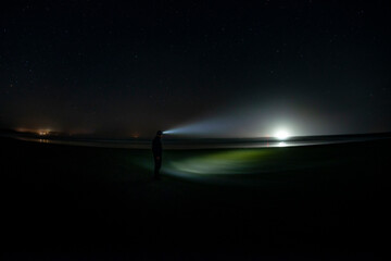 Adventurous man on a beach at night shinning his headlamp at a distant light with stars in the sky. 