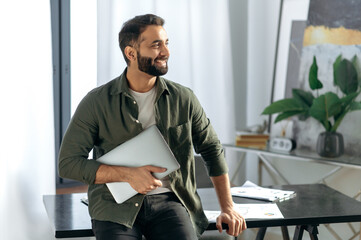 Portrait of an attractive modern Indian or Arabian successful entrepreneur or freelancer in casual clothes, stands in his modern office near the table, holds a laptop, looks away, smiling