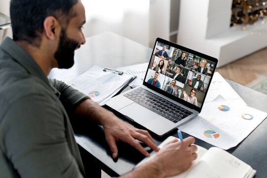 Business briefing. Indian guy looking at laptop screen, talking on video call with multinational business group, holds conference, virtual meeting, brainstorming with colleagues online, takes notes