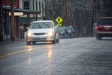 A heavy spring rain comes down in buckets in Windsor in Upstate NY.  A car with headlights on drives down Mainstreet on a wet spring afternoon.