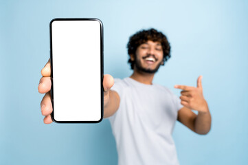 Mock-up, copy space. Defocused indian or arabian guy, in casual wear, holds smartphone with blank white screen for presentation and points finger at it, stands on isolated blue background, smiling