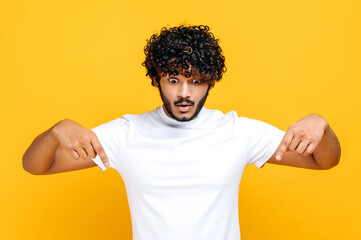 Fototapeta na wymiar Surprised indian or arabian shocked curly-haired guy in basic white t-shirt, amazed looks down and points fingers down, excited face expression, stands on isolated orange color background