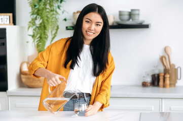 Daily water intake. Positive pretty Chinese girl in casual stylish clothes, standing at home in the...