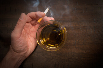 A male caucasian hand holds a smoking cigarette near a glass of whiskey.
