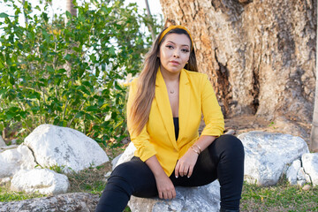 Portrait of a Mexican woman sitting on a stone in a park wearing a yellow jacket.