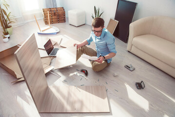 Young man in a blue shirt and trousers is sitting on the floor with a laptop and trying to assemble furniture with his own hands.