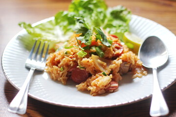 Pork and sausage fried rice served in a white plate on a light wooden table. With lemon, vegetables and tomato slices on a light wooden table.