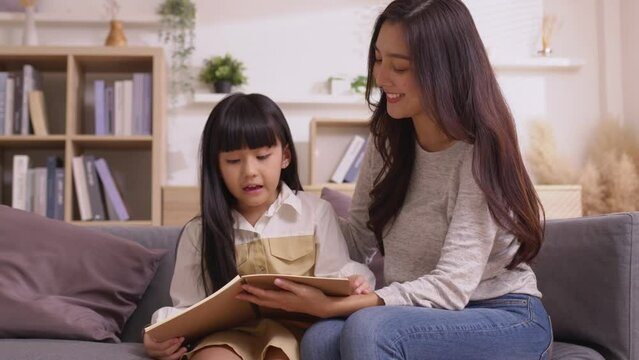 Asian young mother smiling reading book to cute teen daughter sitting on couch in relax day.