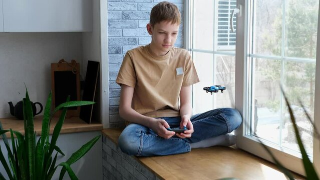 Blond boy launches toy drone at home. Teenager sits on windowsill and plays a drone