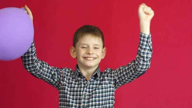 Joyful caucasian boy 7-8 years old with a balloon rejoices and raises his hands up. Studio shooting, red background. The child rejoices at his birthday with a balloon