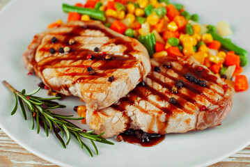Pieces of meat grilled. Steak with rosemary and pepper in a white plate. Meat dish with red sauce on a white wooden table. Salad of corn, carrots and green peas near the steak.