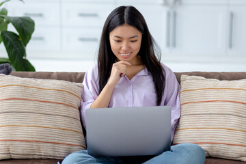 Portrait of happy Asian woman using laptop to browse social media or funny video while sitting on sofa. Relax at home concept