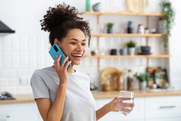 Smiling African American woman holding glass of clean water, talking on mobile phone while standing in kitchen at home, healthy lifestyle concept