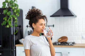 Healthy lifestyle concept. Beautiful young Afro American woman drinking clean water standing at home in the kitchen, copy space