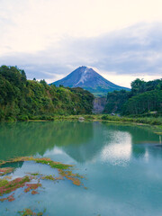 The view of Mount Merapi and a lake with a greenish water and rocky surface, the sky looks cloudy. Forest vegetation that looks still dense with trees surrounding the lake. Bego Pendem, Mount Merapi