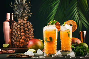 Alcoholic cocktail with vodka, pineapple juice, mango, liquor, ice. Long drink or summer mocktail. Tropical dark background with palm leaves and exotic fruits - obrazy, fototapety, plakaty