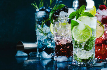 Cold summer cocktails drinks. Classic alcoholic long drink and mojito or mocktail in highballs with blueberries, blackberries, raspberries, lime, herbs and ice on blue background