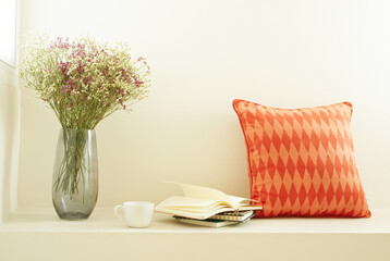 Dried pink and white Statice flower with glass vase with book and coffee cup under warm sunlight beside window and orange geometric pattern pillow on beige background , relaxation corner concept