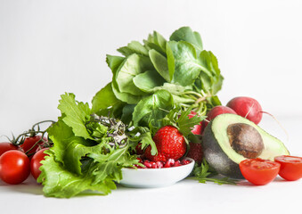 Various salad ingredients: green lettuce leaves, avocado, tomato, pomegranate seeds and strawberries on table at white wall background. Healthy food lifestyle with vitamin rich food. Front view.