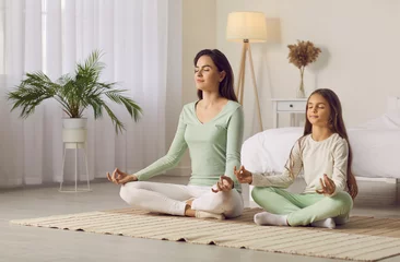 Foto op Canvas Family yoga. Beautiful young woman and her charming little daughter are smiling while doing yoga together at home. Family sits in lotus position on floor in living room. Mom teaches child to meditate. © Studio Romantic