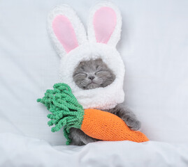 Funny kitten wearing easter rabbits ears sleeps with knitted carrot on a bed under warm white...