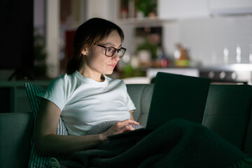 Serious young woman in glasses using laptop sitting on couch at home in the night. Thoughtful girl freelancer watching education video, lying on sofa and working on computer in dark room. 