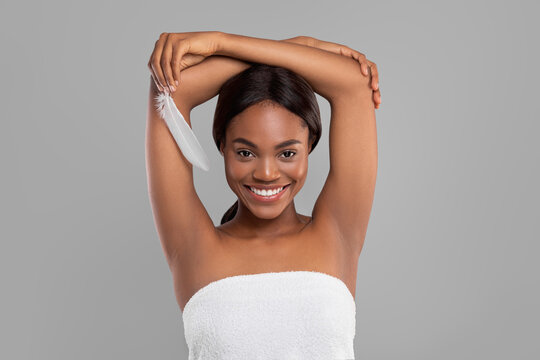 Smiling pretty millennial black female with perfect skin showing armpits and hold feather, isolated on gray background