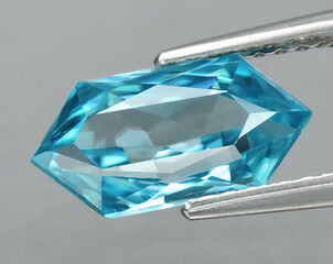 Natural gemstone blue zircon in tongs on a gray background