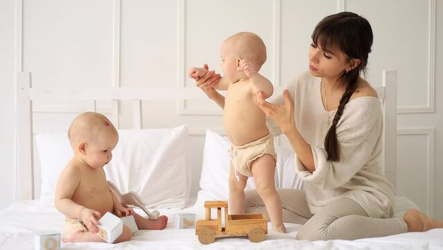 mom with twin babies in diapers playing with wooden toys at home on a bed in a bright white room, maternal love and care with two small children