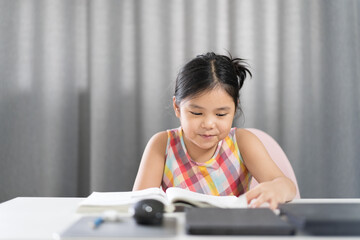 Asian child student or kid girl smile reading on book to learning and study online class with computer laptop on desk or person happy enjoy learn from home to read for back to school by homeschool
