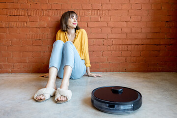 Young woman sitting relaxed on floor while robotic vacuum cleaner cleaning floor at home. Concept of smart home gadgets and comfort