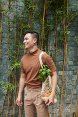 Portrait of happy young man holding mesh bag of fresh groceries he bought at local market