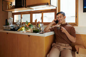 Smiling young man sitting at kitchen counter, drinking morning coffee and talking on phone with friend