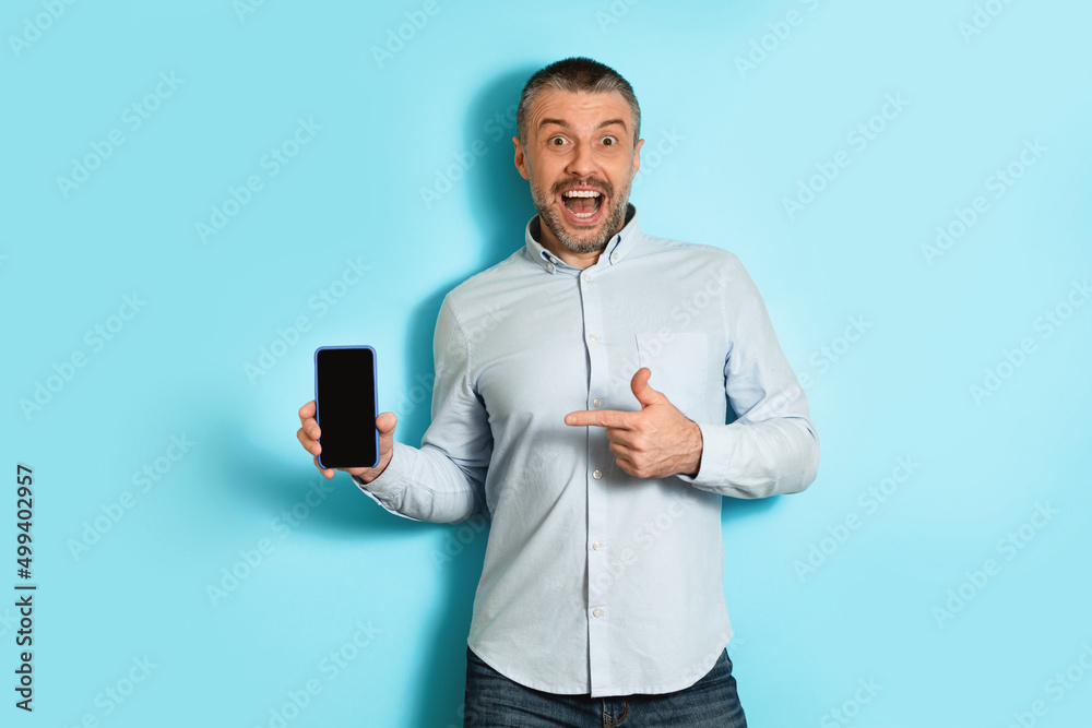 Poster excited man showing smartphone empty screen pointing finger, blue background