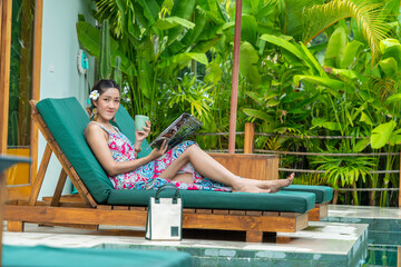 Asian tourists, Asian tourists, wearing dresses, lounging in the sun loungers by the pool, reading magazines during the summer vacation. - obrazy, fototapety, plakaty