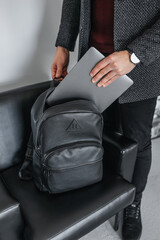 A man in a jacket puts his hand on a gray laptop in a stylish black leather backpack.
