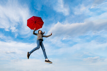 Little happy girl fly on background of blue cloudy sky holding red umbrella. Imagination, adventure, weather forecast, windy weather concept.