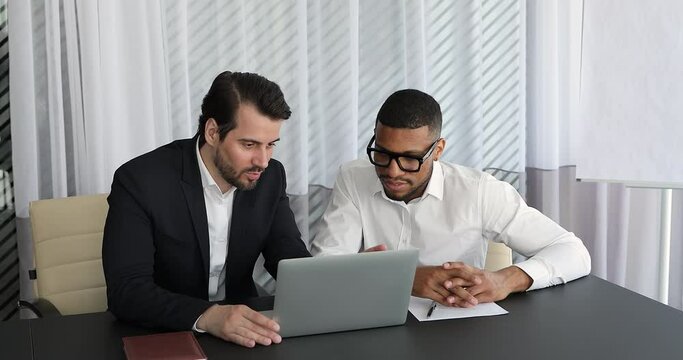 Experienced confident male mentor sit at desk with Black man intern trainee teach younger colleague to prepare report in electronic app on laptop. Leader coach help new worker explain job on workplace